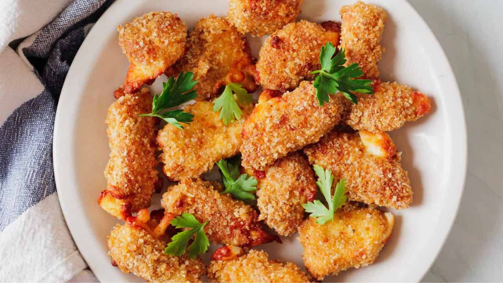 A plate of breaded chicken nuggets garnished with parsley. The nuggets are golden brown, and the dish is placed on a white surface with a striped cloth partially visible.