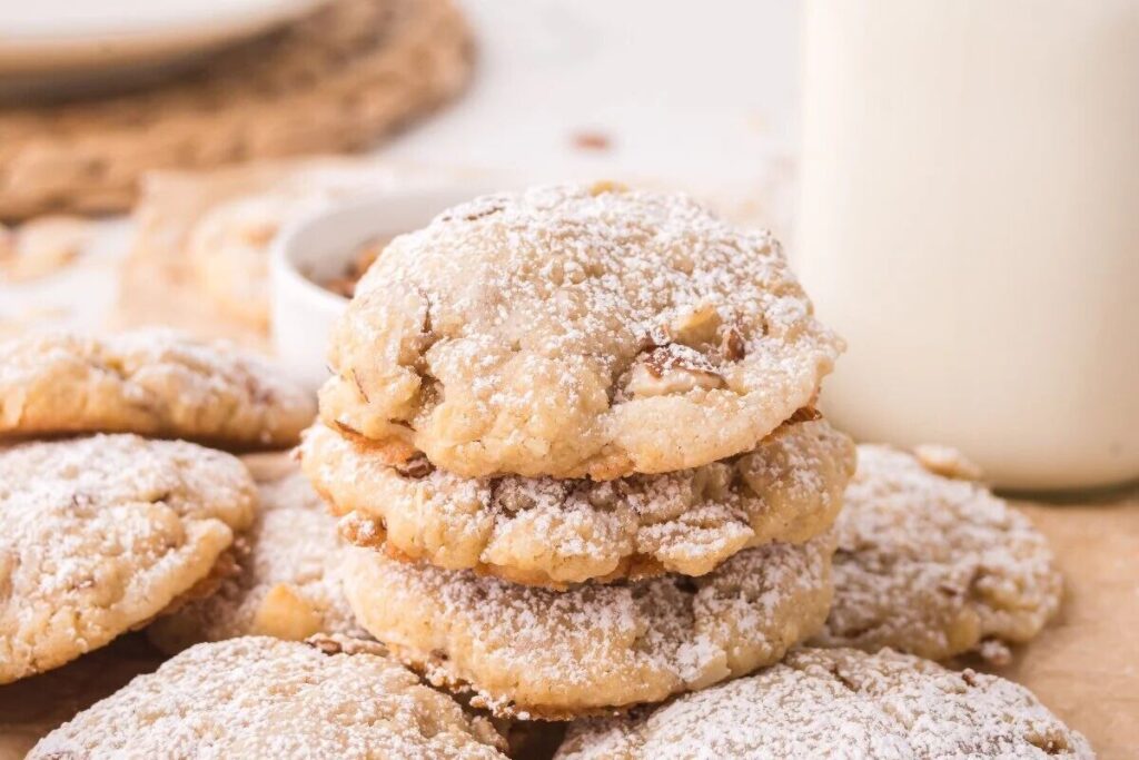 A stack of powdered Almond Croissant Cookies sits next to a glass of milk on a light surface.