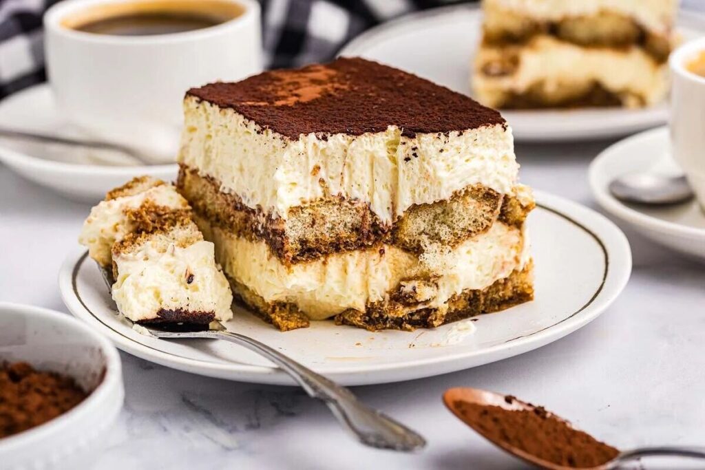 A slice of Italian tiramisu sits on a white plate, layered with creamy goodness and dusted with cocoa, with a spoonful ready to enjoy.