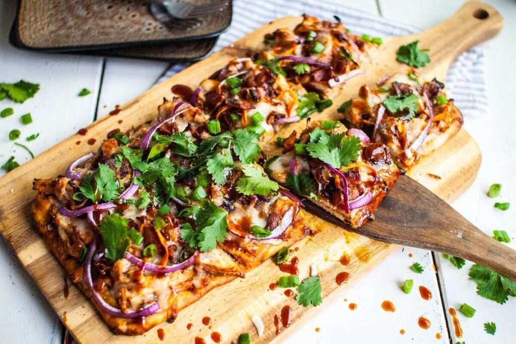 Rectangular pizza with red onions, cilantro, and sauce on a wooden board with one slice being served. Enjoy easy BBQ chicken pizza inspired by Panera at home.