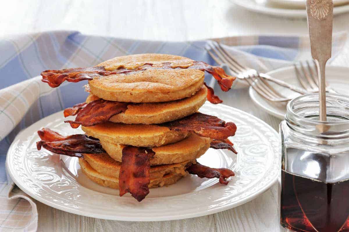 A stack of pancakes with strips of bacon, being drizzled with syrup from a spoon, on a white plate with a fork beside it.