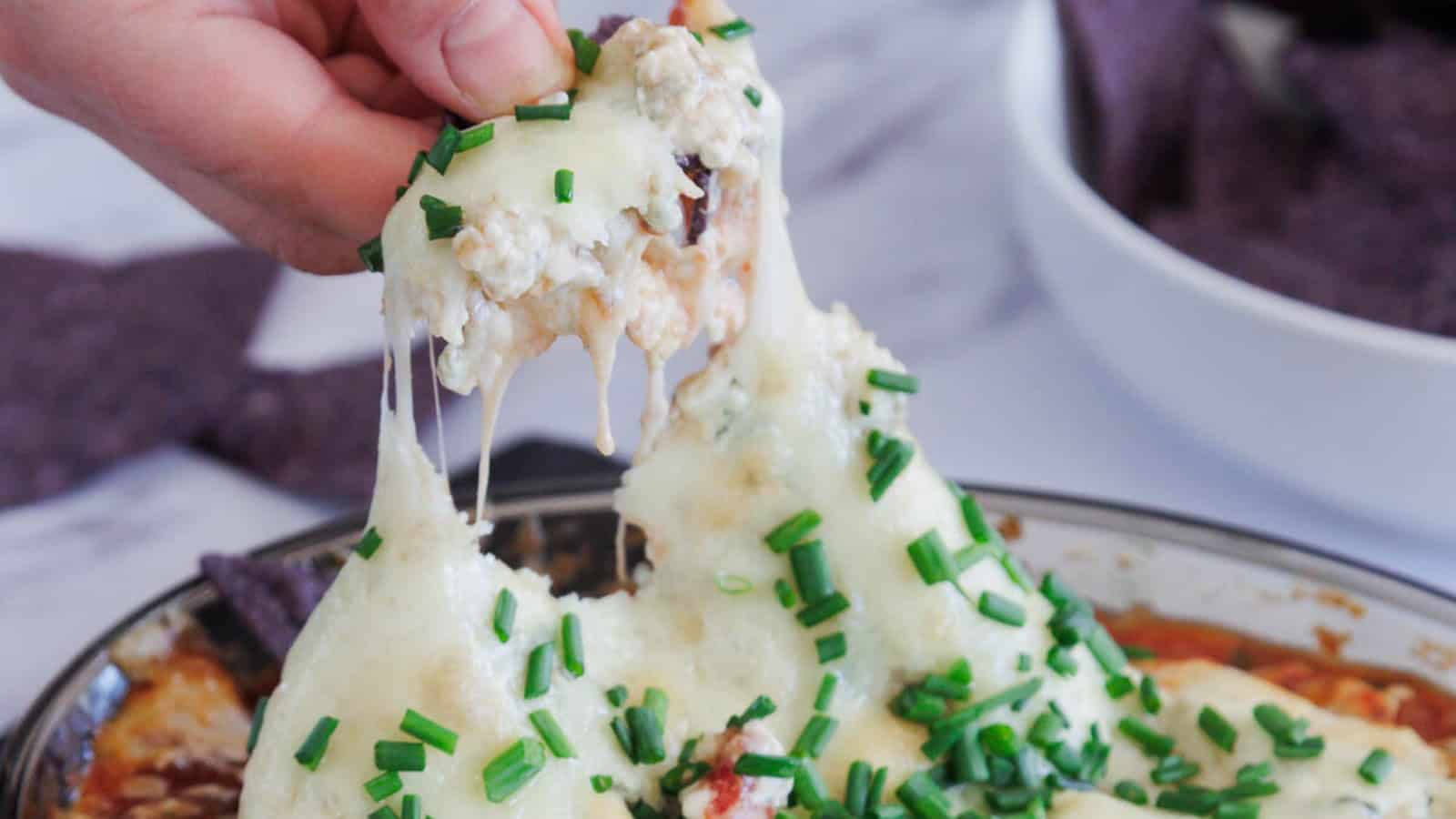 A round glass dish filled with a baked cheesy casserole topped with chopped green chives. Two triangular blue corn tortilla chips are inserted upright on one side of the dish.
