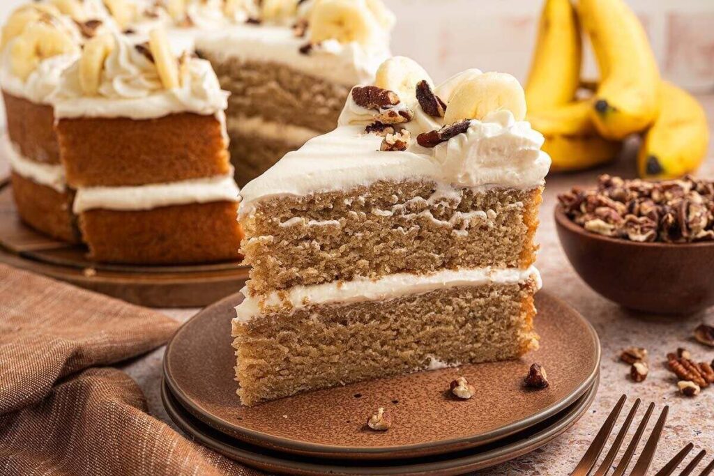 A slice of banana cake with cream frosting, served with fresh bananas and nuts.
