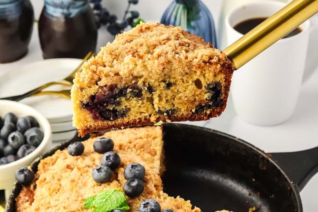 A slice of blueberry crumb cake is lifted from a cast-iron skillet, with coffee and fresh blueberries in the background.