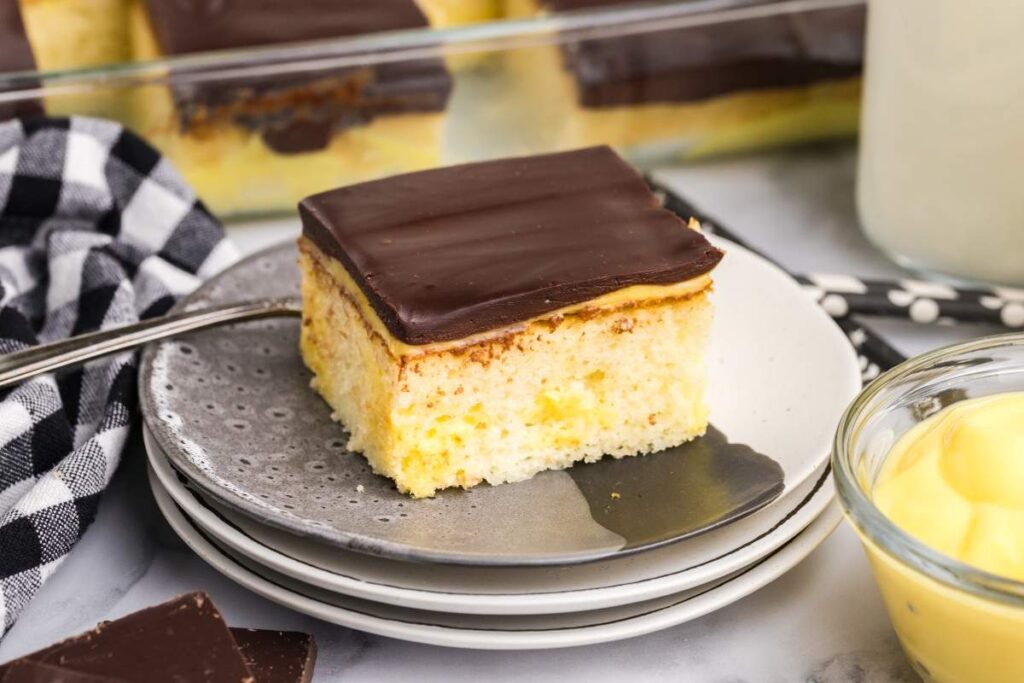 A slice of Boston cream poke cake on stacked plates with a checked napkin and pudding bowl.