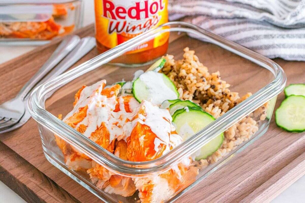 A glass container with buffalo chicken, crisp cucumber, and fluffy rice, with hot sauce in the background.