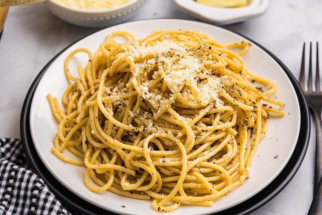 A plate of Cacio e Pepe glistens under the light, the cheese and black pepper blending perfectly into the pasta. The checkered napkin and polished fork create a welcoming scene. It is a dish that calls for slow enjoyment and pure delight.