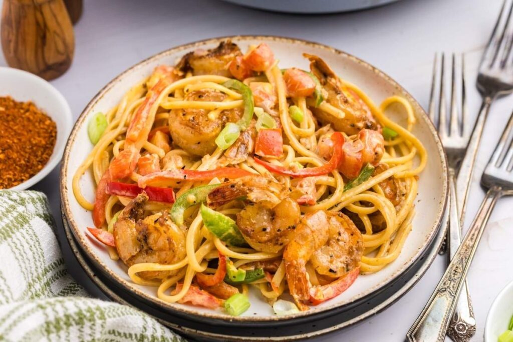 A plate of Cajun shrimp pasta featuring succulent shrimp, vibrant red bell peppers, and crisp green onions, accompanied by forks and a napkin.