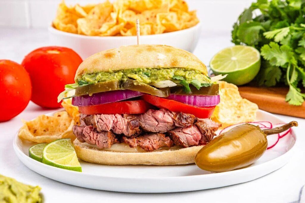 Juicy carne asada torta with steak, tomatoes, onions, jalapeños, and guacamole, served with lime and chips.