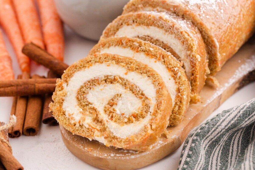 Sliced carrot cake roll with cream filling, served on a wooden board with cinnamon sticks and fresh carrots.