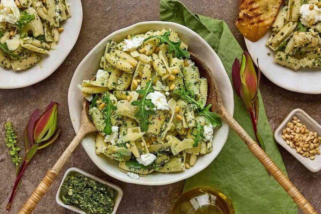 A bowl of cavatelli with arugula, pesto, ricotta, and pine nuts sits with small bowls of pesto, pine nuts, and artichoke buds.