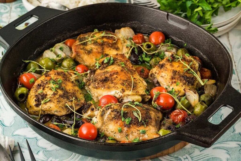 Roasted chicken thighs with herbs in a black skillet, surrounded by cherry tomatoes, olives, and onions. 