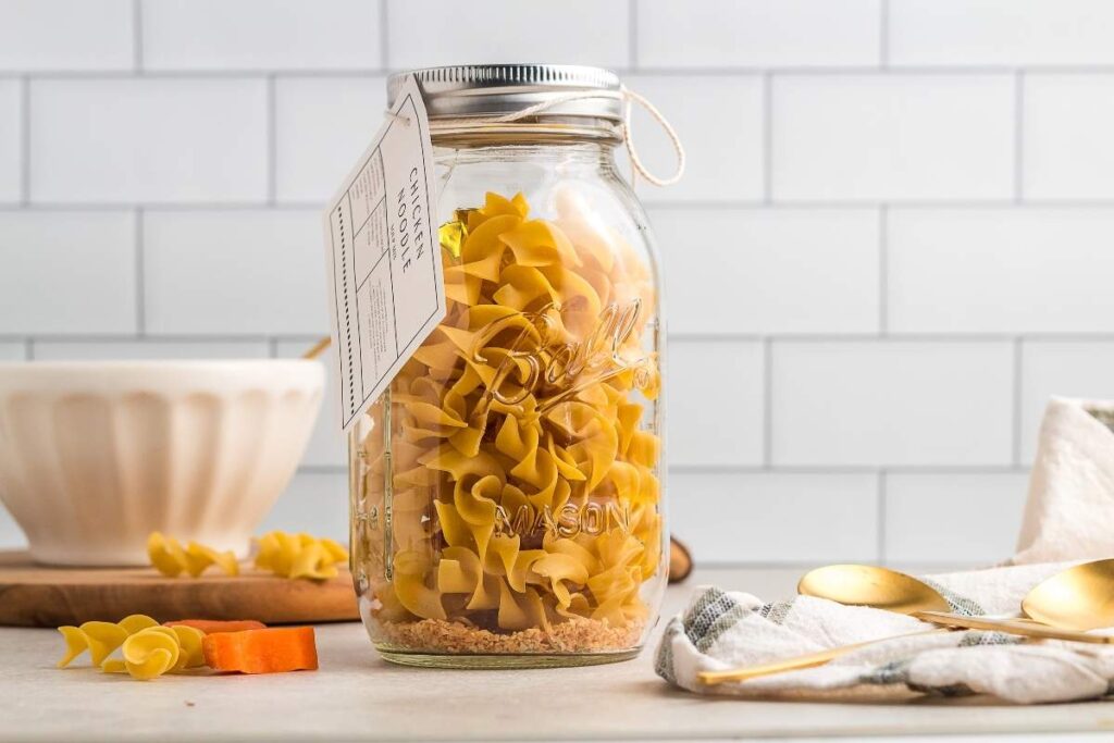 A mason jar filled with pasta and labeled "chicken noodle soup in a jar" sits on a countertop, with a recipe card attached.