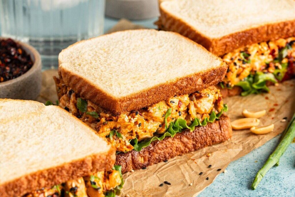 Two Chicken Salad Sandwiches with Chili Crisp on brown parchment paper, next to a small bowl of dark sauce and a glass of water in the background.