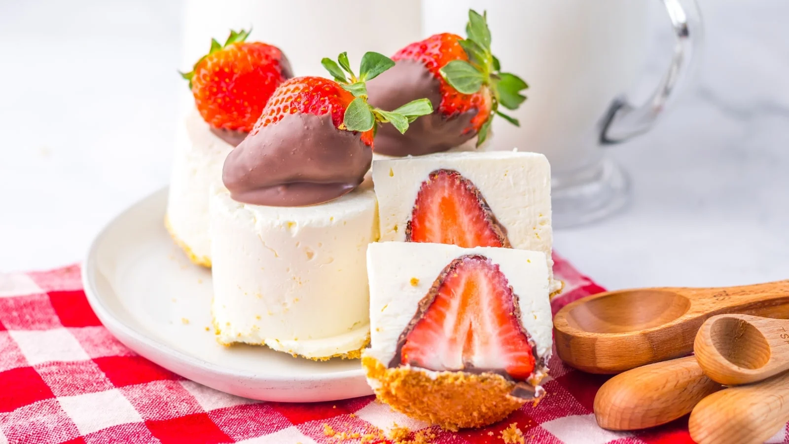 Cheesecake slices topped with chocolate-covered strawberries on a white plate. Red checkered cloth and wooden spoons beside.