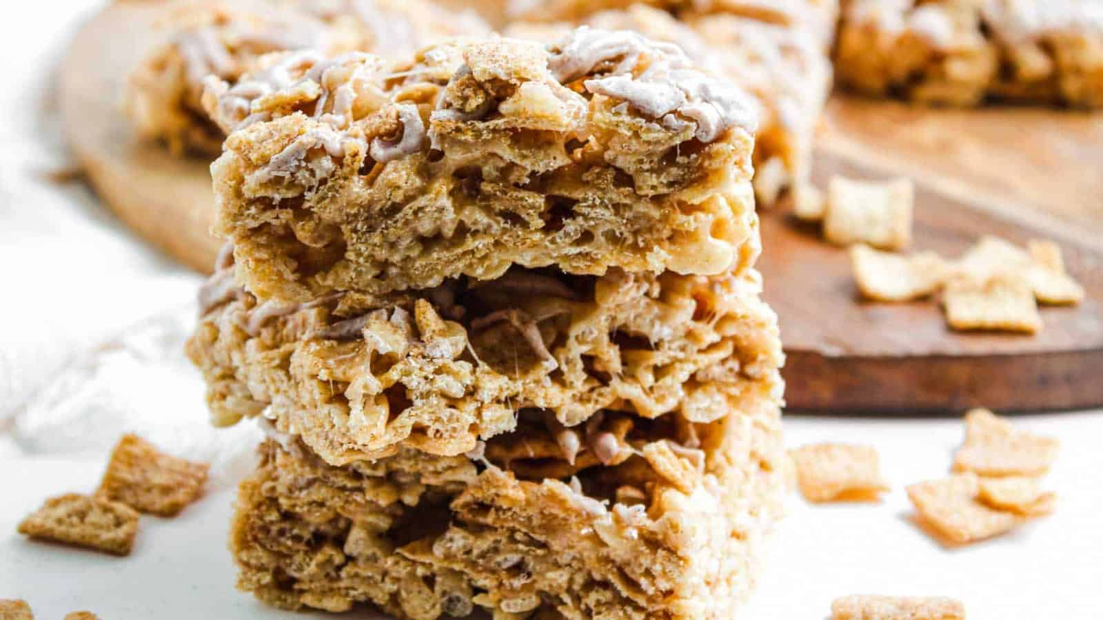 Three cinnamon cereal bars stacked, with cereal pieces scattered around on a white surface and wooden board.