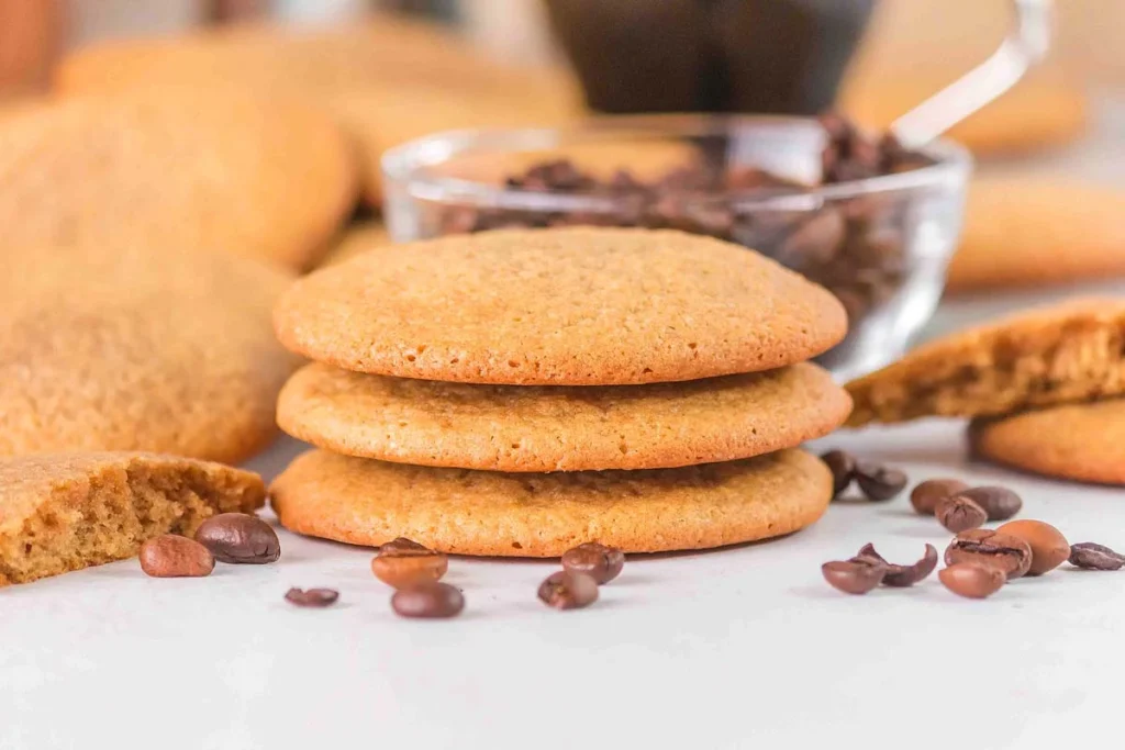A stack of three coffee cookies sits among scattered coffee beans, with a bowl of beans and a spoon in the background.