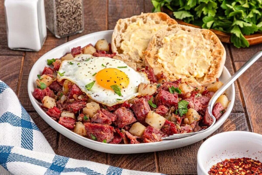 Plate of corned beef hash with diced potatoes and a sunny-side-up egg, served with buttered English muffins.