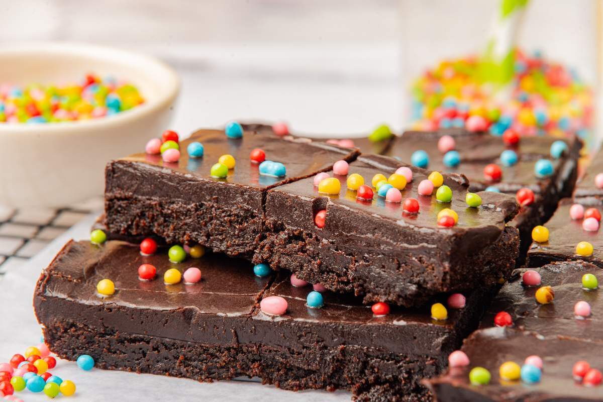 Close-up of a thick, fudgy chocolate brownie topped with colorful candy sprinkles on a light blue surface.