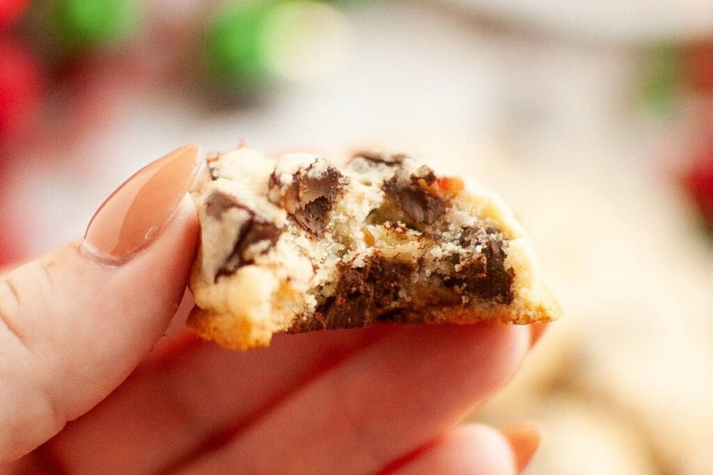 A half-eaten chocolate chip cookie held between fingers with a peach-toned manicure, showing a cross-section revealing a soft, light-colored interior speckled with melted chocolate chunks and a golden-brown, slightly crisp edge.