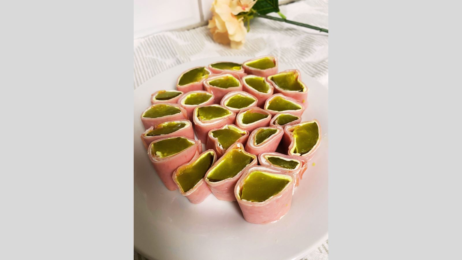 Ham slices rolled and filled with pickles, arranged on a white plate. A pale flower and part of a green stem are blurred in the background.