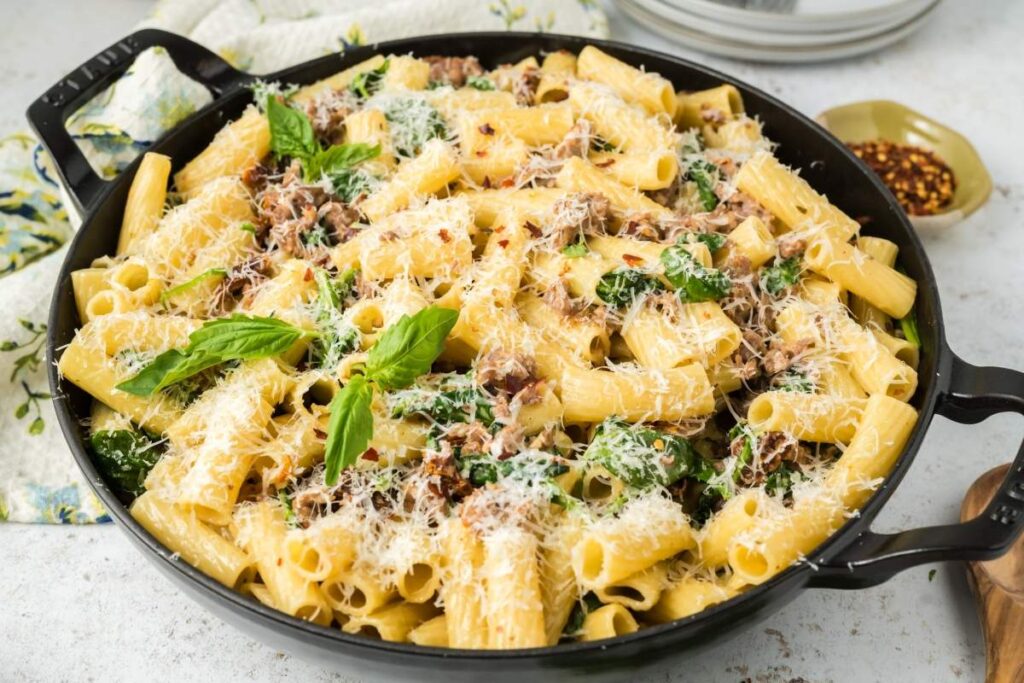 A skillet filled with creamy Tuscan sausage pasta, mixed with shredded meat, spinach, and topped with grated cheese and basil leaves.