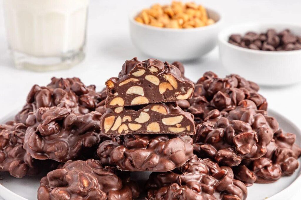Crockpot Candy chocolate clusters with peanuts sit on a plate, surrounded by bowls of chocolate, peanuts, and a glass of milk.