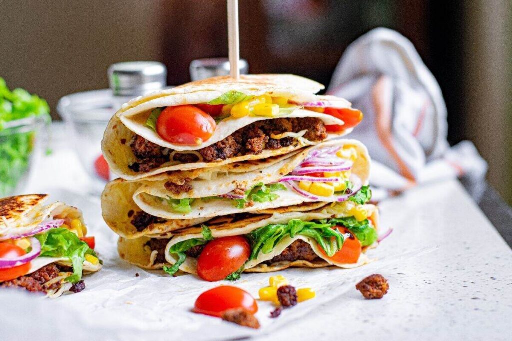 Three stacked tacos with ground meat, lettuce, tomatoes, onions, and corn on a countertop.