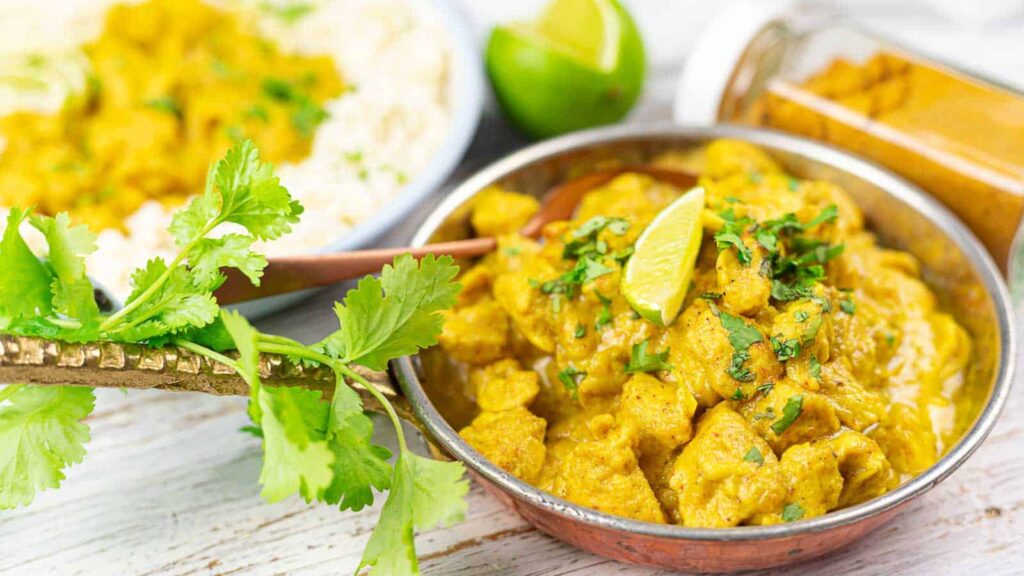 A bowl of chicken curry topped with cilantro and a lime wedge, with a jar of turmeric and a plate of rice in the background.