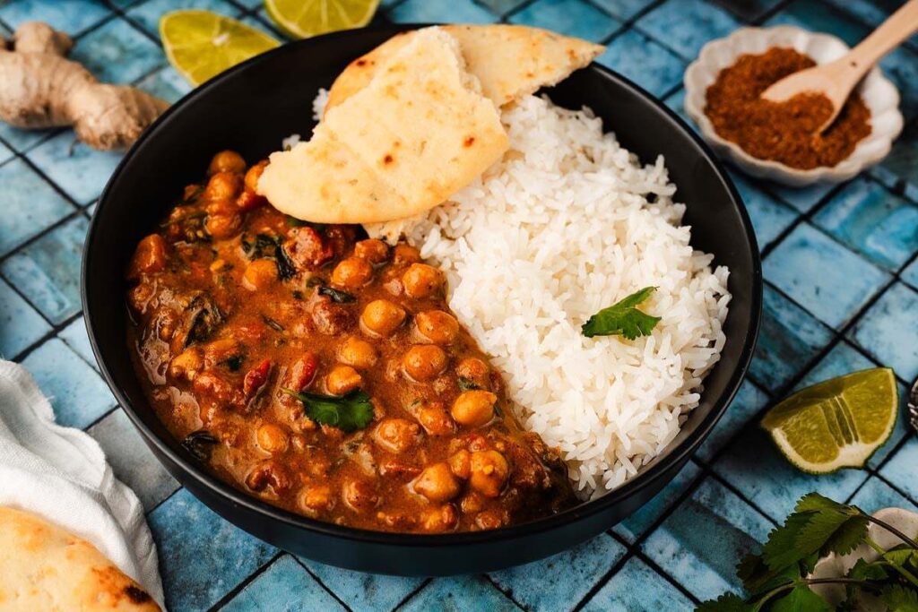 A black bowl of chickpea curry with spinach sits with white rice and naan, garnished with cilantro.