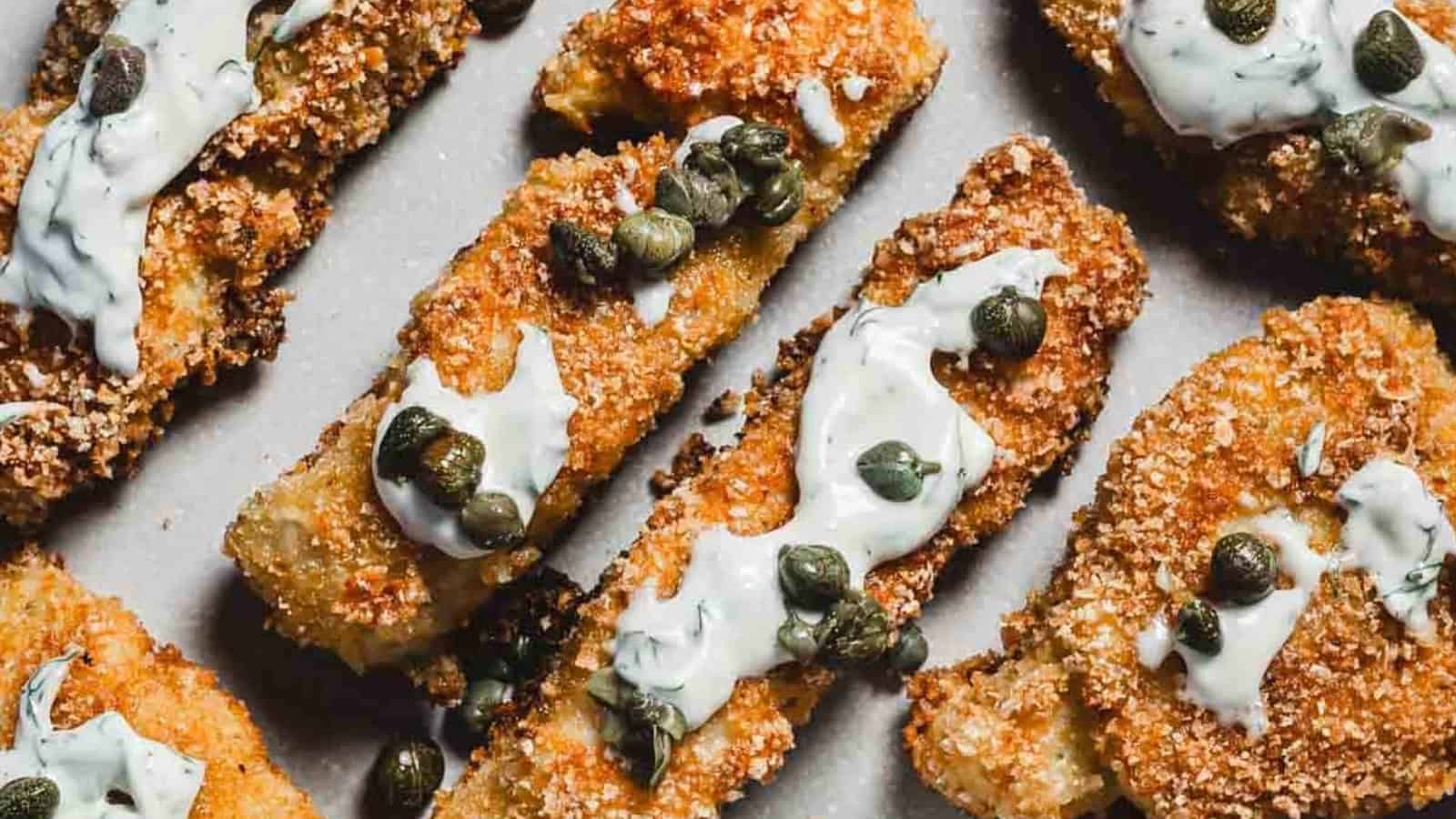 Golden breaded fish sticks topped with creamy sauce and capers, arranged on a light-colored surface.