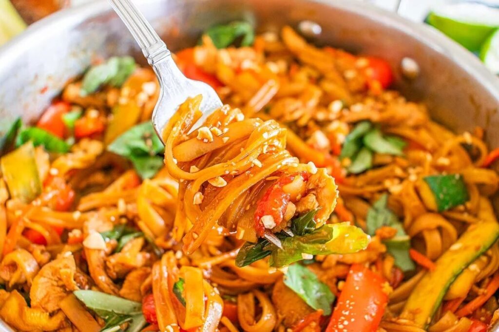 A fork holding a portion of Thai drunken noodles with vegetables, chicken, and garnished with peanuts, hovers above a pan brimming with the same delicious dish.