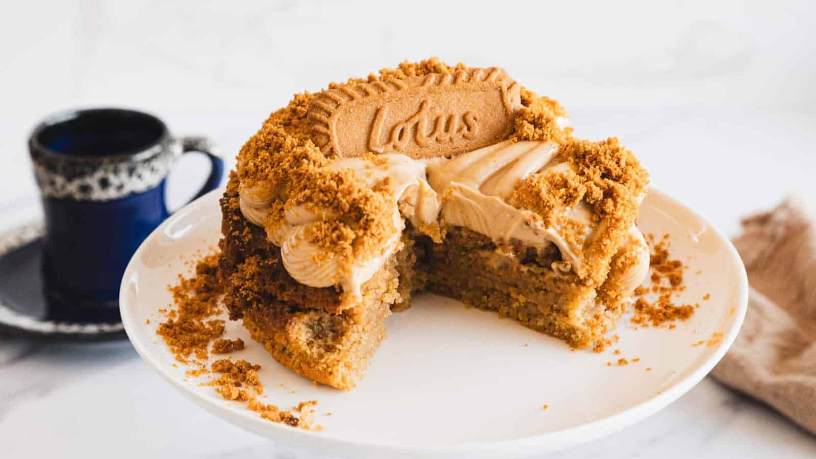 A partially sliced cake with light brown frosting and crumbled Lotus Biscoff cookies on top, displayed on a white cake stand. A blue cup sits in the background.