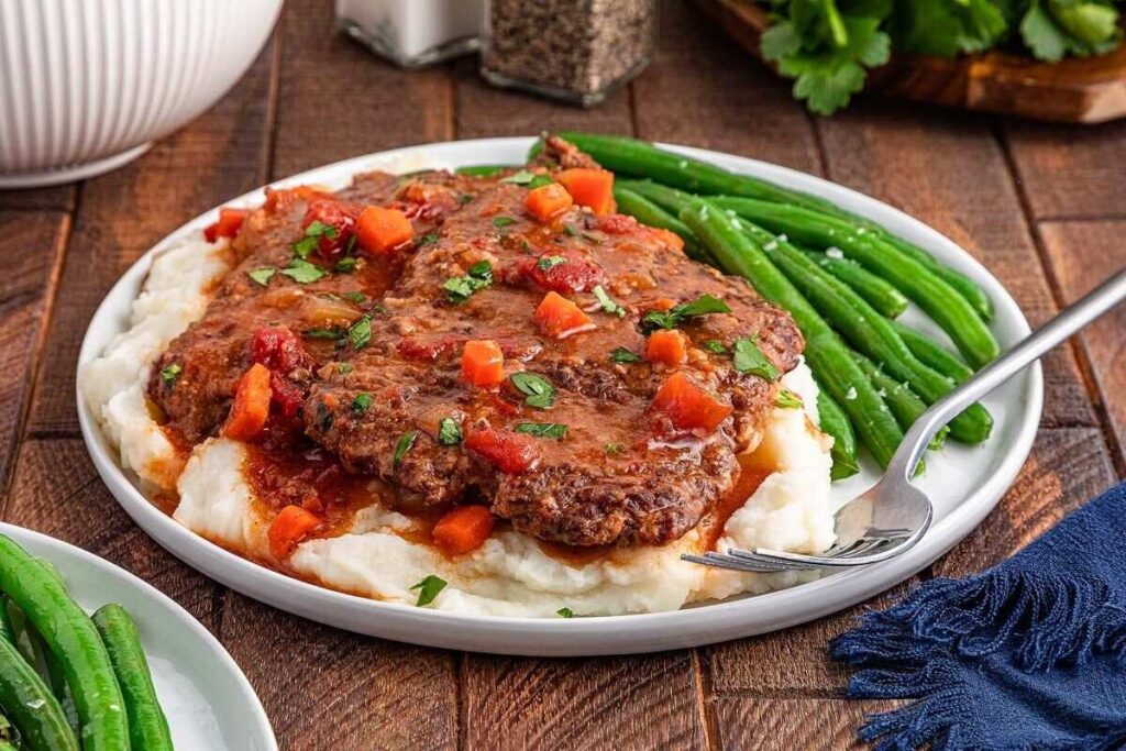Swiss steaks with gravy and vegetables over mashed potatoes, served with green beans and a fork.