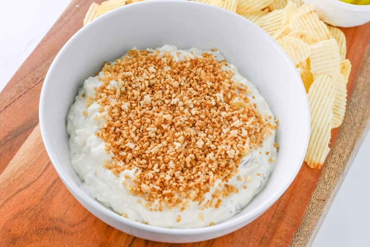 A bowl of creamy dip topped with crumbled breadcrumbs sits on a wooden board, surrounded by wavy potato chips.