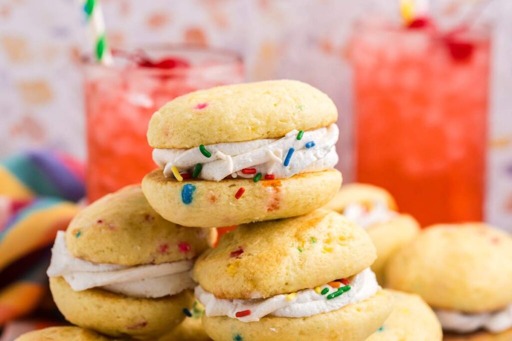 A stack of funfetti cake mix whoopie pies with sprinkles and white frosting filling with two red beverages in the background.
