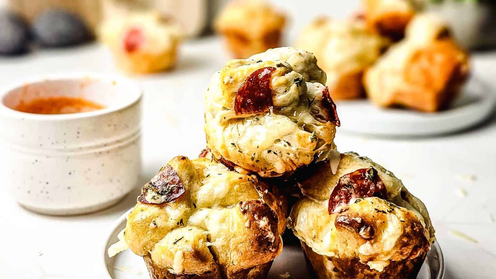Three muffin-shaped pieces of pull-apart bread with melted cheese and pepperoni are stacked on a plate. A small bowl of dip and more bread pieces are visible in the background.