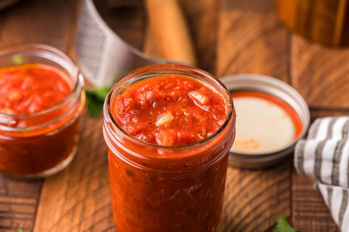 A glass jar filled with tomato sauce sits on a plate. Two spoons are beside it. A small bowl is in the background, slightly out of focus.