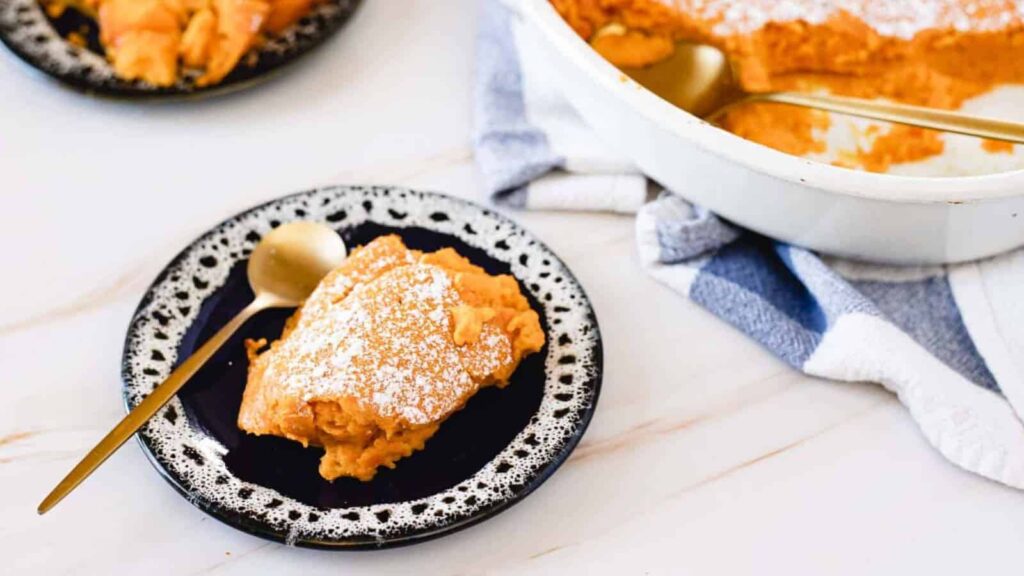 A slice of Carrot Soufflé on a black and white plate. In the background is a casserole dish with more of the dessert and a white and blue towel.