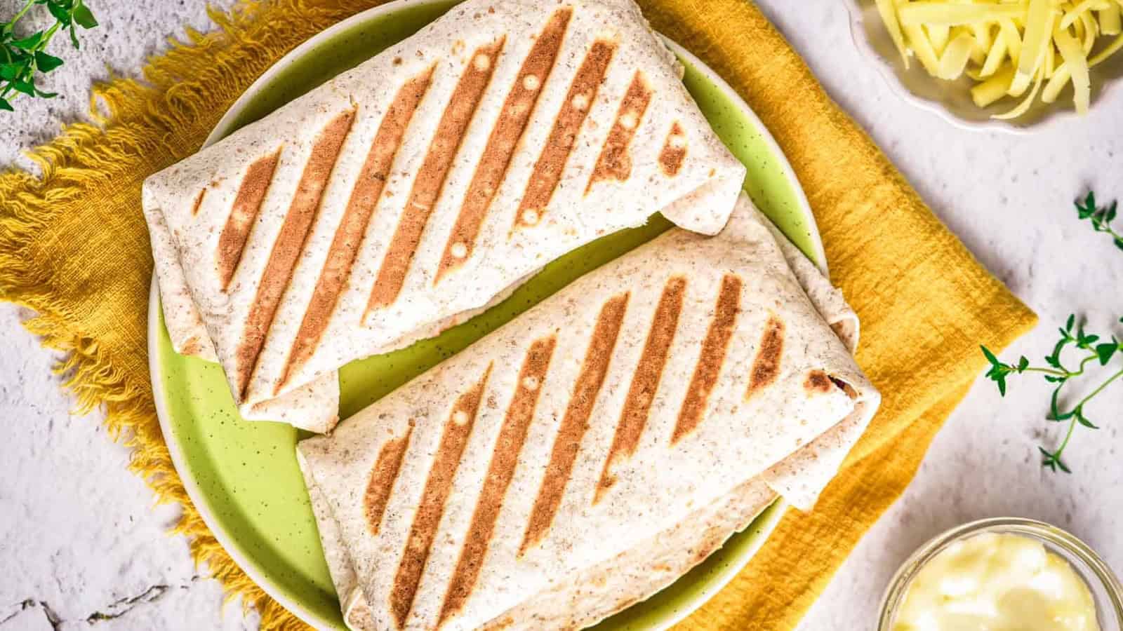 Two grilled wraps with visible grill marks are placed on a green plate with a yellow cloth underneath. Shredded cheese and a small bowl of sauce are seen nearby on a white surface.