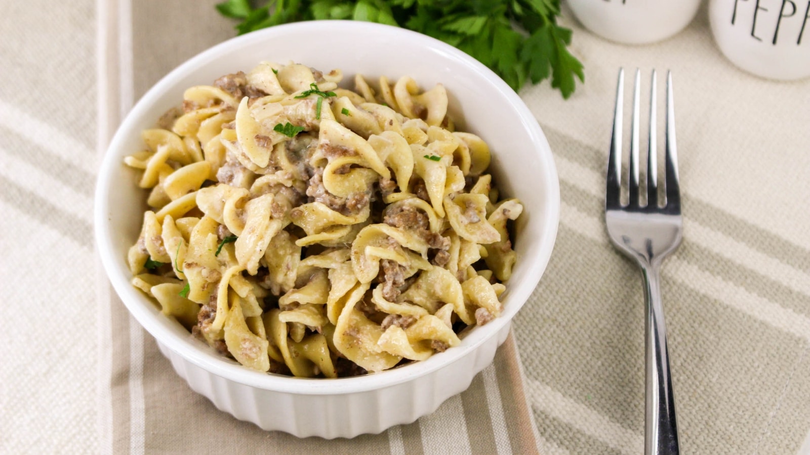 A bowl of creamy beef stroganoff with egg noodles on a striped cloth next to a fork and a sprig of parsley.