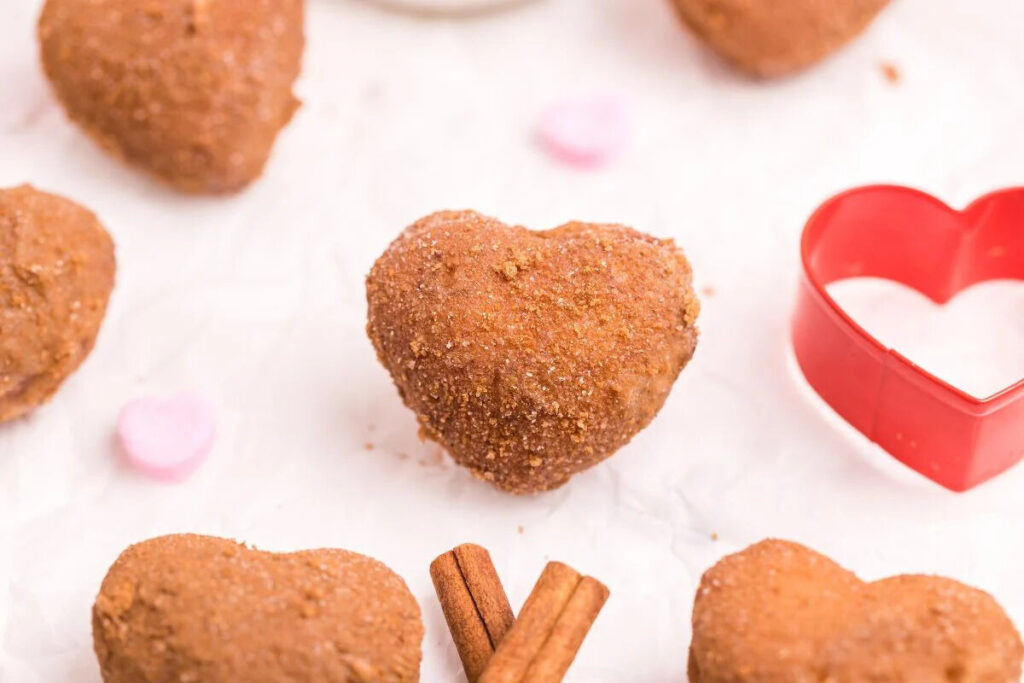 Heart-shaped donut coated in cinnamon sugar, surrounded by candy hearts and a red heart-shaped cookie cutter.
