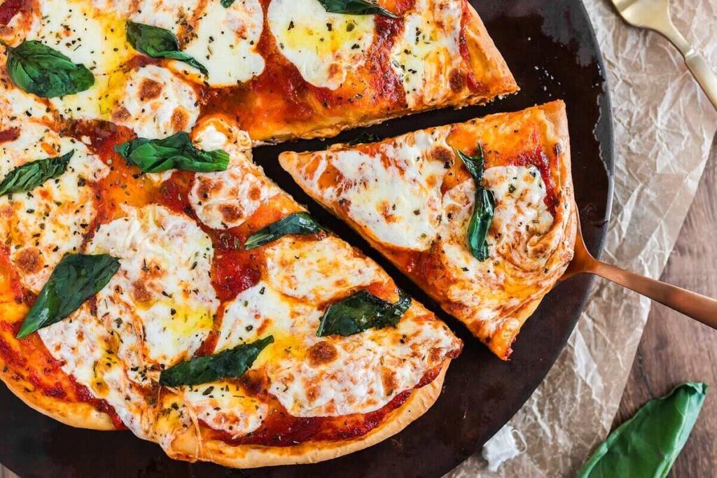 A margherita pizza with fresh basil and melted mozzarella cheese is sliced, with one delicious slice being lifted on a spatula.