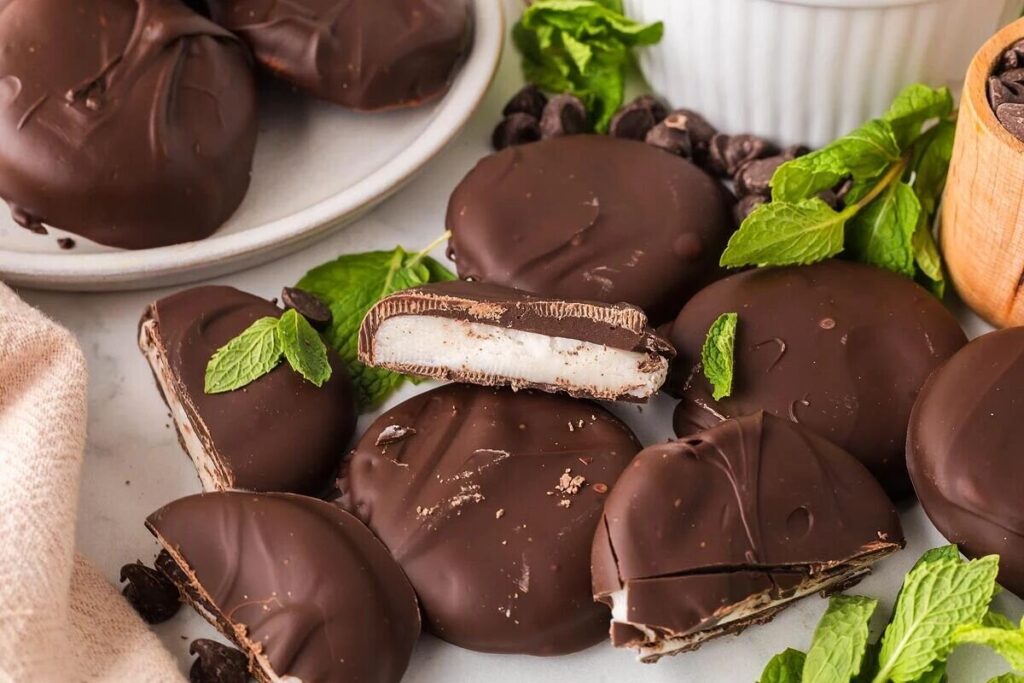 Homemade York peppermint patties sit on the table, some whole and some sliced, with mint leaves and chocolate chips around them.