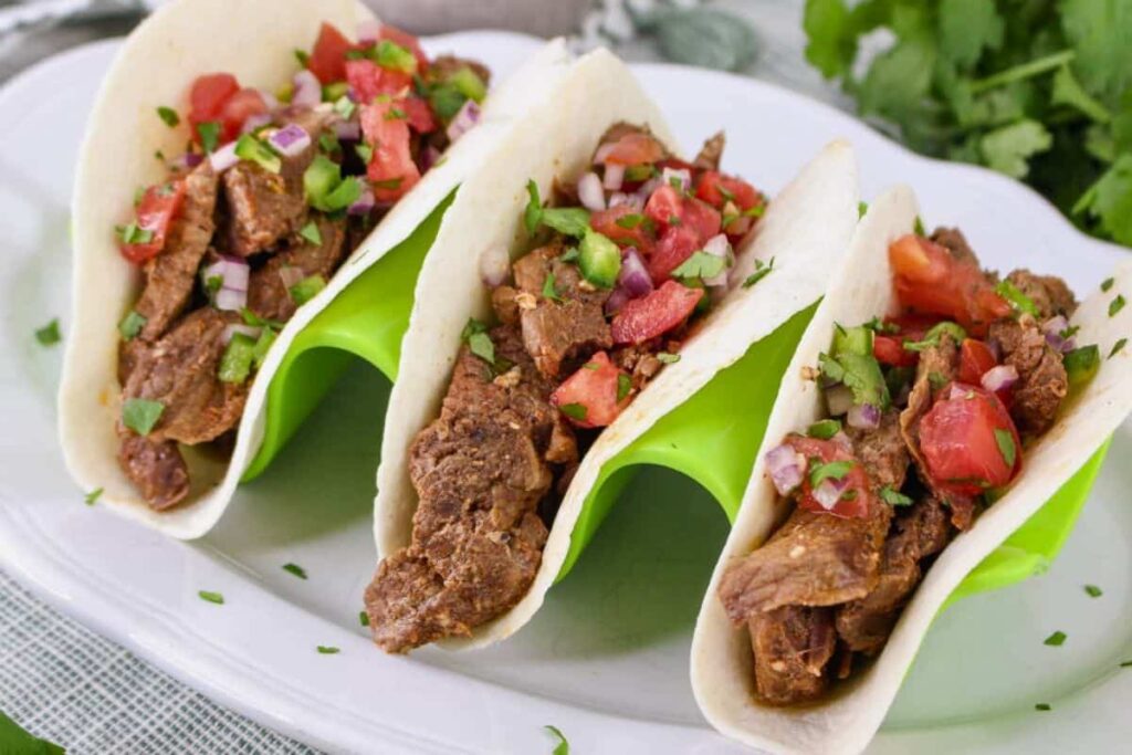 Three beef tacos with diced tomatoes, onions, and cilantro served in soft tortillas on a white plate with a green holder.