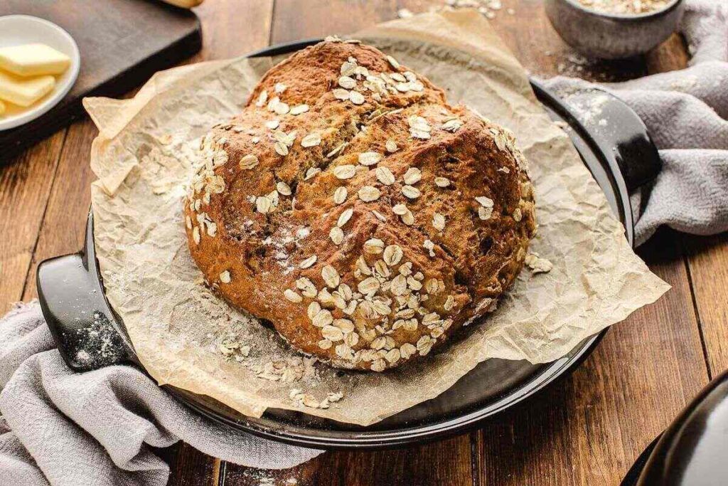 A loaf of oat-topped Irish soda bread rests on parchment paper on a dark round tray. 