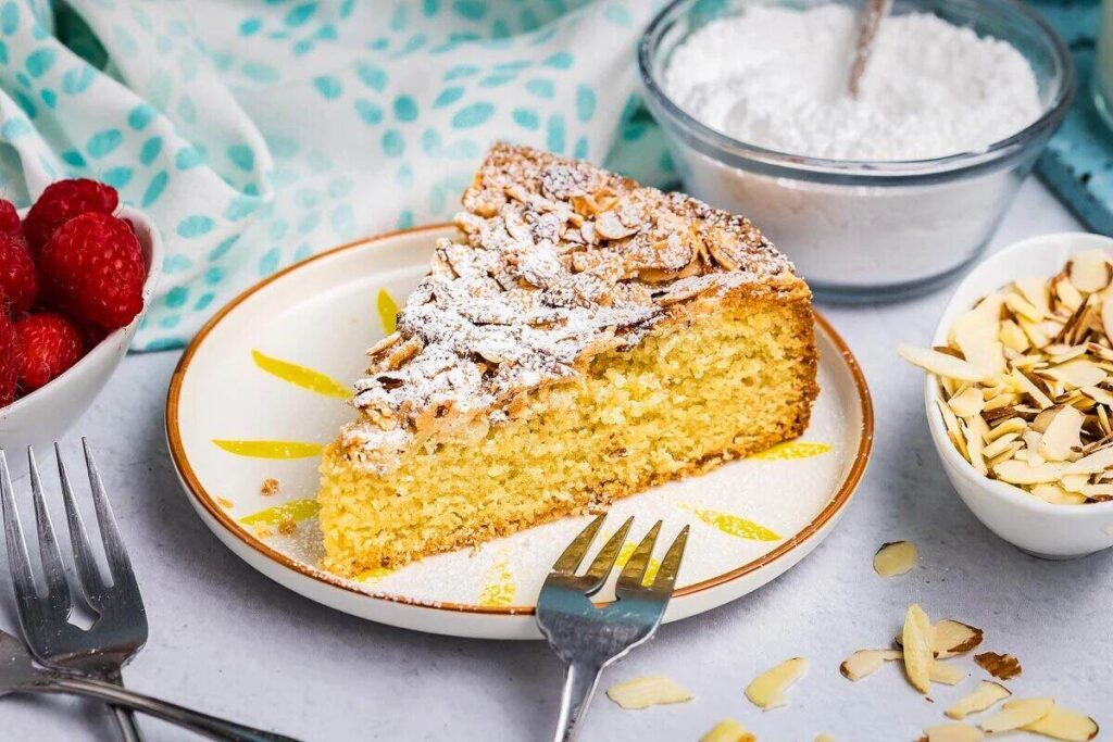 A slice of Italian almond cake dusted with powdered sugar, served with raspberries, sliced almonds, and a fork.