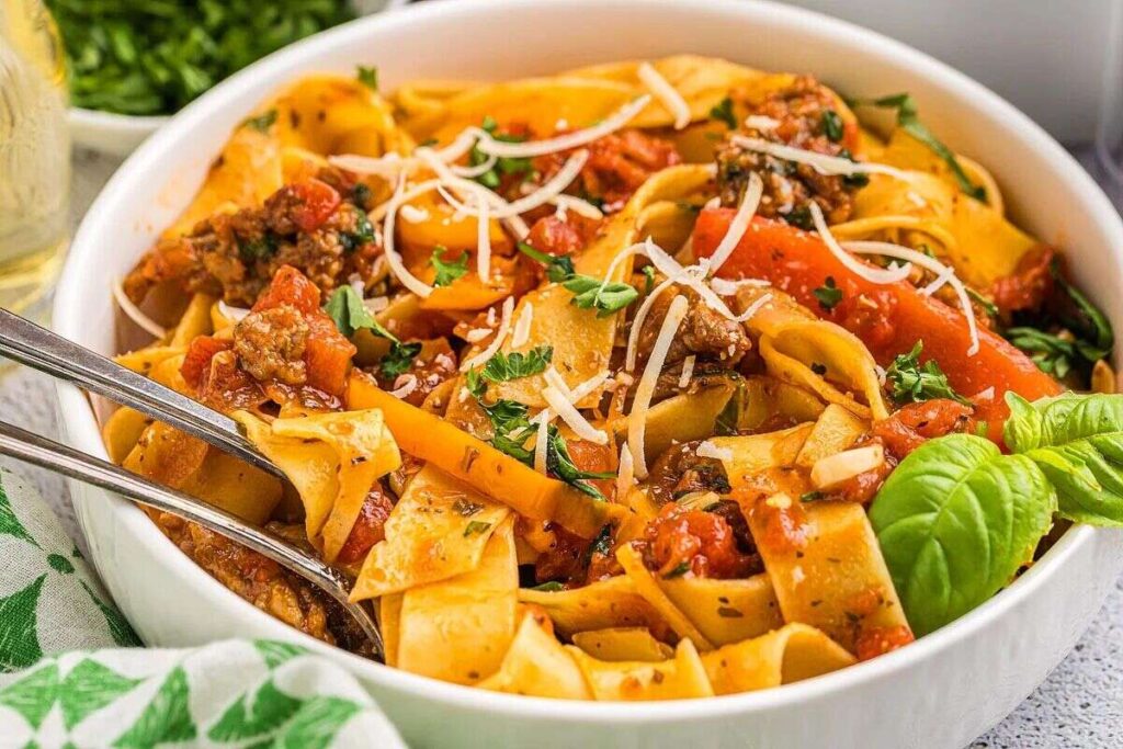 A bowl of Italian drunken noodles, topped with ground meat, tomatoes, shredded cheese, and garnished with basil leaves.