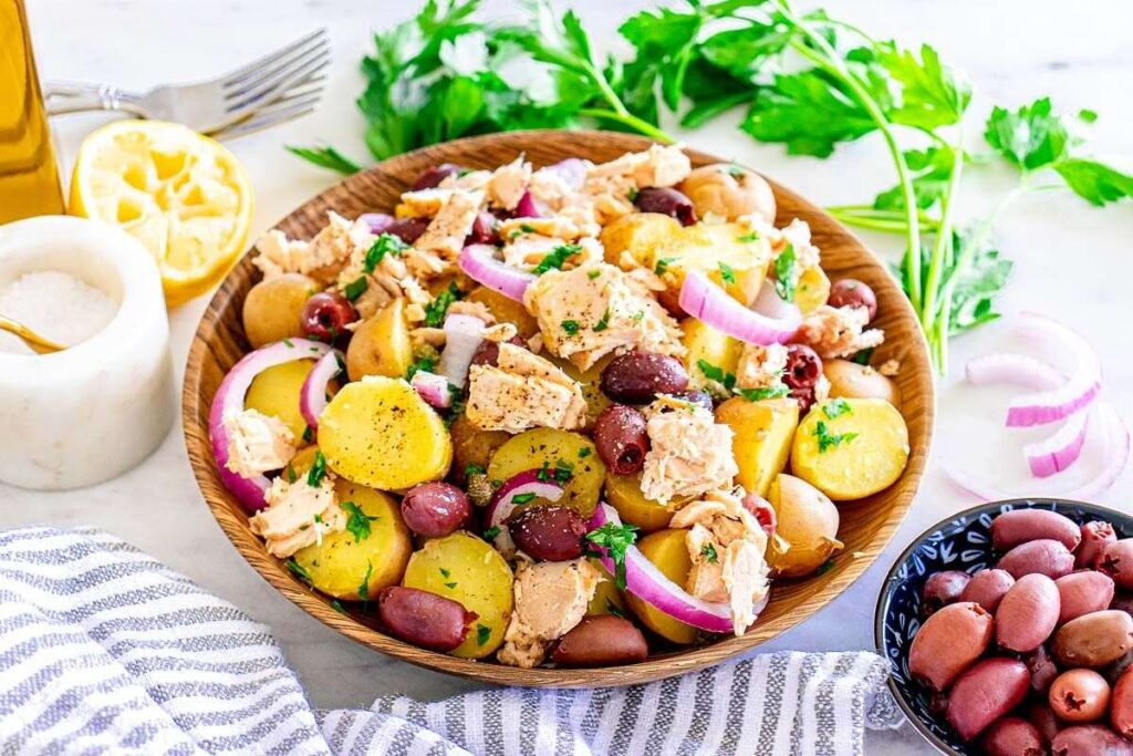 Italian tuna potato salad with red onions, olives, and parsley, served with a half lemon, extra olives, parsley sprigs, and a salt cellar on a white surface.
