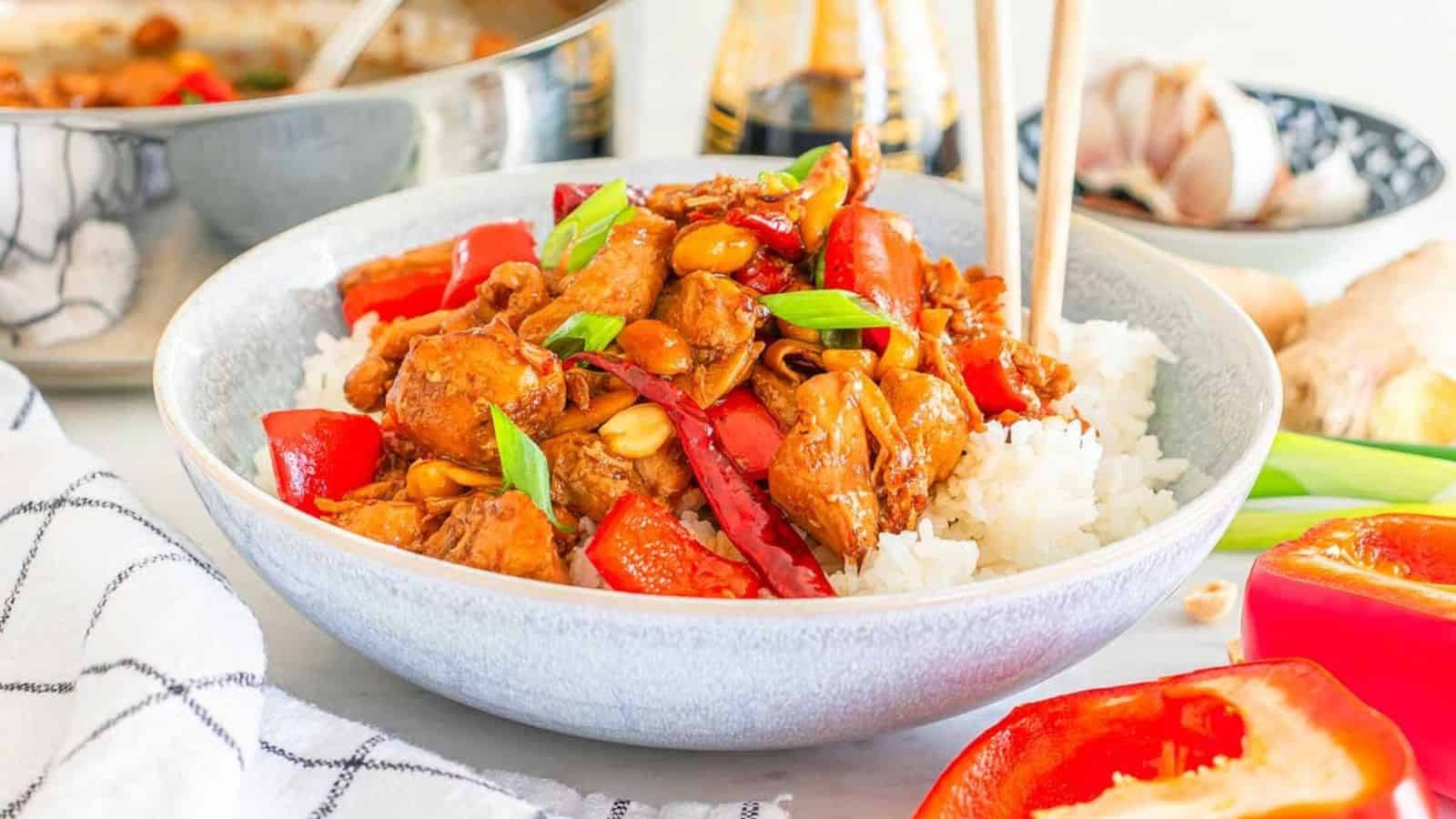 Bowl of stir-fried chicken with red bell peppers, peanuts, and green onions on white rice, next to chopsticks and cooking ingredients.